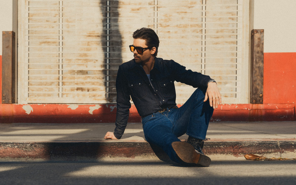Man sitting on a curb wearing sunglasses and a dark jacket with a red and beige wall in the background.