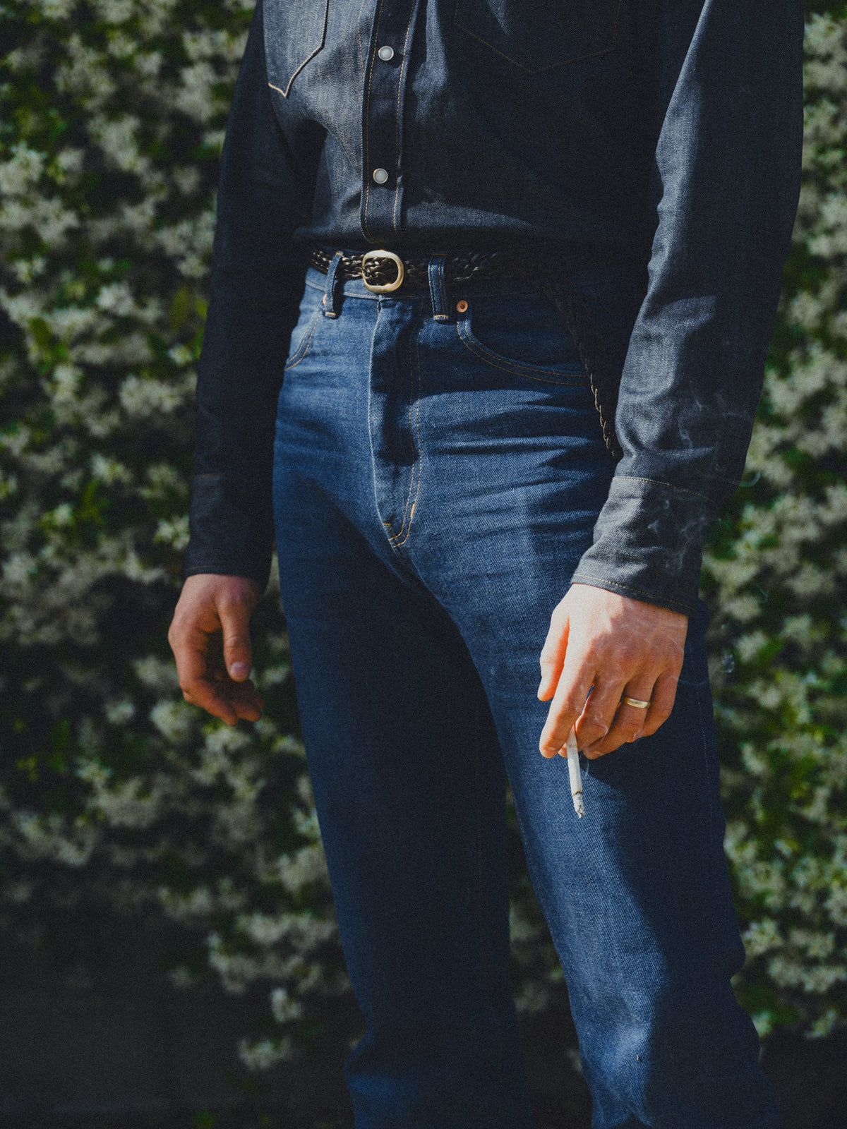A person wearing dark blue denim jeans and a matching dark denim long-sleeve shirt, standing outdoors against a blurred green hedge background.