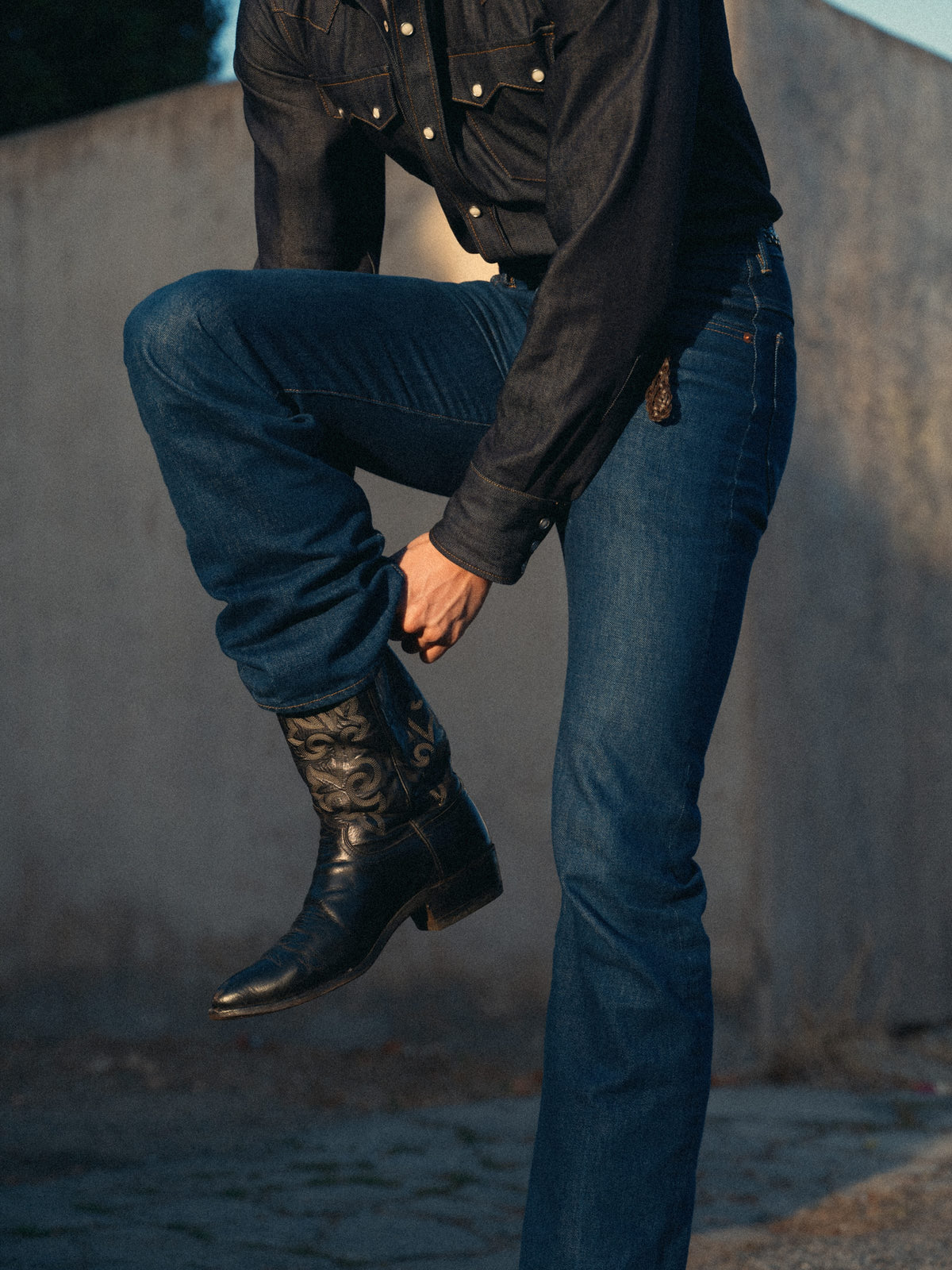 A person wearing dark blue jeans and black western cowboy boots with embroidered detailing, posed outdoors against a textured concrete wall.