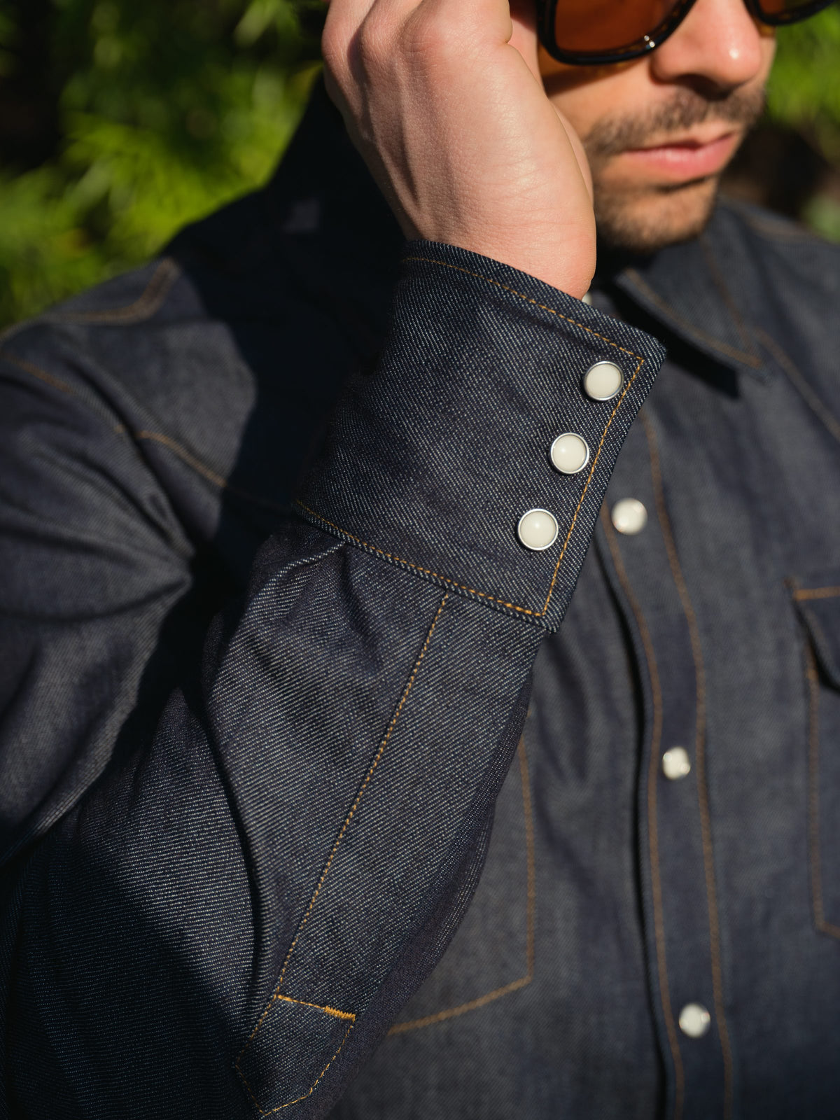 Close-up of a man wearing a dark indigo denim shirt jacket with white buttons and contrast tan stitching, outdoors with green foliage blurred in the background.