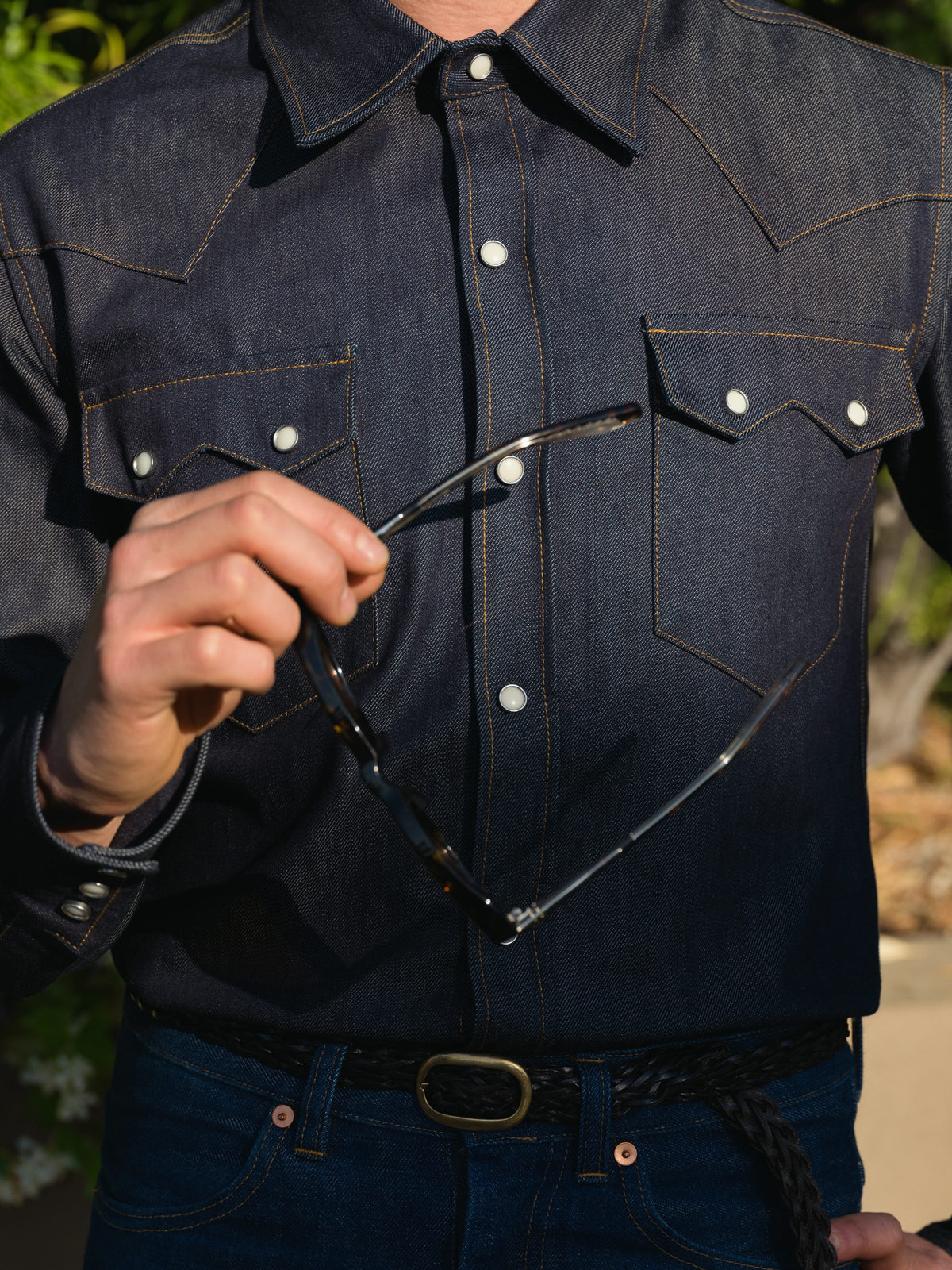 A close-up of a person wearing a dark indigo denim western shirt with contrast gold stitching and white snap buttons, holding a pair of sunglasses in one hand.