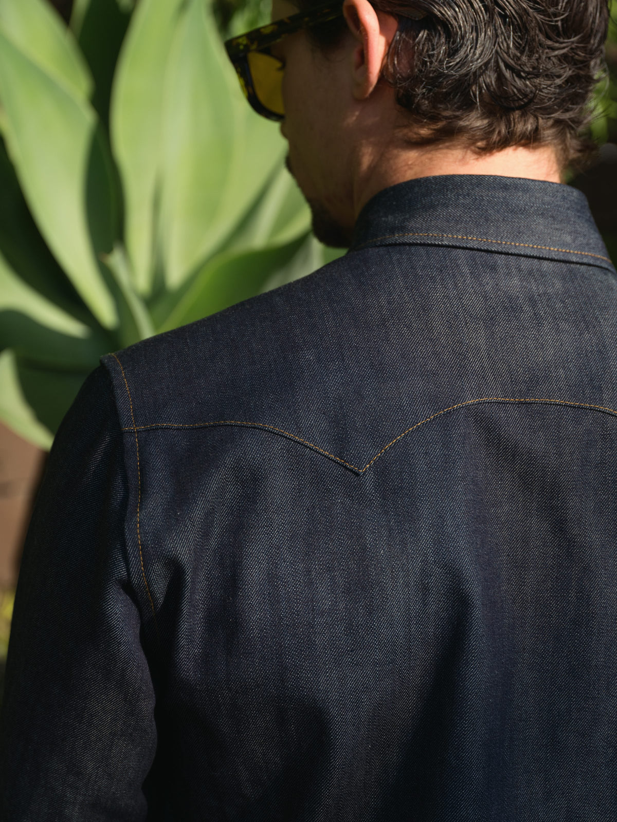 A close-up back view of a man wearing a dark indigo denim shirt with contrast tan stitching, set against a background of green foliage.
